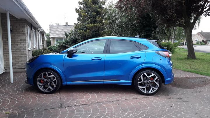 RE: 2022 VW T-Roc R vs. Ford Puma ST Gold Edition - Page 2 - General Gassing - PistonHeads UK - The image shows a blue hatchback parked in front of a house. The car is positioned at an angle, highlighting its sporty design with black and red wheels and a large spoiler on the rear wing. It features a sunroof and tinted windows, adding to its sleek appearance. The vehicle also has several visible modifications such as lowering kit and aftermarket rims, indicating it may be used for performance driving or racing. The surroundings suggest a quiet residential area with well-maintained lawns and a clear sky above.