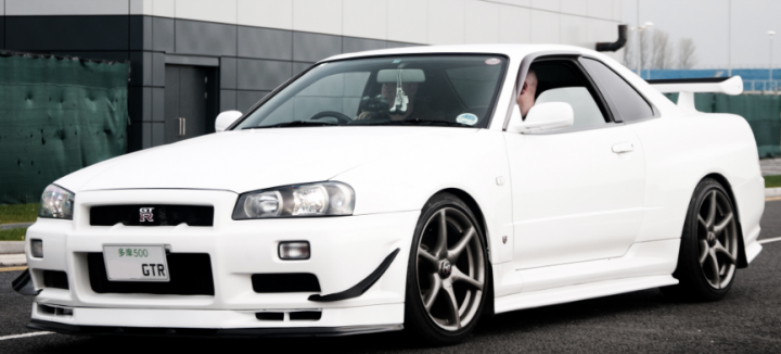 A white car parked in a parking lot - Pistonheads - The image depicts a white sports car parked outdoors. The car's bodywork appears to be in pristine condition, with a shiny and smooth finish, signifying attention to detail. It has distinctive headlights and alloy wheels, which are indicative of a thriving custom scene. A license plate is visible at the front of the car. The car is in a stationary position, with its driver exiting through the driver's side door. The backdrop reveals an industrial environment with a warehouse or a building in the distance.