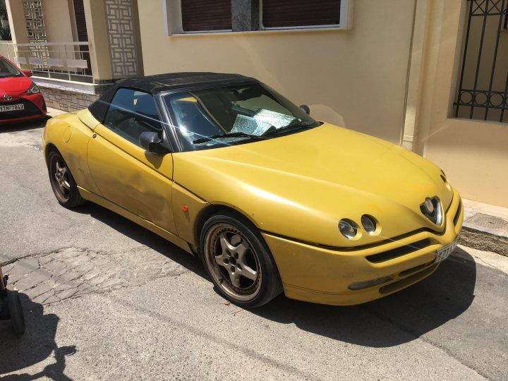 Let's see your Alfa Romeos! - Page 114 - Alfa Romeo, Fiat & Lancia - PistonHeads - The image shows a vibrant yellow sports car parked on the side of a street. The car is a convertible with its top down, revealing its interior. It has a sleek design typical of high-end models, and it's positioned at an angle that provides a clear view of its front and side profile. In the background, there are residential buildings, suggesting this could be a suburban or urban area. A few personal items are visible near the car, including what appears to be a pair of sandals and a handbag, possibly belonging to the car's owner.