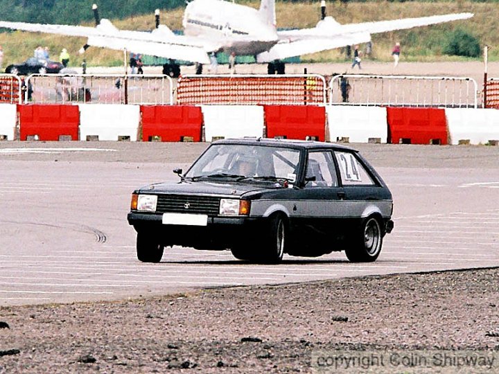 Specials Worst Pistonheads Homologation - The image captures a dynamic scene at an airport runway. A small black car with distinctive orange headlights is driving on the tarmac, positioned in the foreground. The runway is marked by white and red stripes, leading the viewer's eye towards a white airplane parked in the background. The background also includes a barrier and a few bystanders, adding to the sense of a bustling airport setting. The photograph appears to be taken during the day, with clear visibility of the car and the airplane.