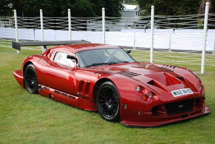 TVR Cerbera Speed 12 pictures. - Page 1 - Goodwood Events - PistonHeads - The image showcases a vibrant red sports car parked on grass, with a backdrop of a fence and white buildings. The car is sleek with a large rear wing, which is a design feature that adds aerodynamic efficiency. In front of the car, there's a small plant, indicating the car has been posed in the outdoor environment. The car appears to be well-maintained, with a glossy finish that reflects the surrounding greenery.