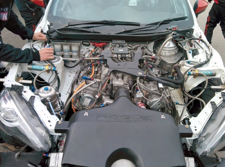 May Bank Holiday at Castle Combe - Page 1 - South West - PistonHeads - The image shows a scene of mechanics working on the engine of a white sports car. The car is parked with the hood opened, revealing the complexity of its internal mechanics. The mechanics are focused on the engine, with one person at the top of the engine bay, and another leaning in from the side. Various tools and equipment are scattered around the engine, including metal wrenches and several small bottles, suggesting a repair or maintenance process is in progress. The setting appears to be an outdoor parking area, with the hood propped open to allow easy access to the engine.