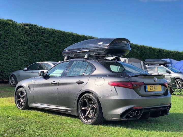 Alfa Romeo Roof Bars - Giulia QV - Page 1 - Alfa Romeo, Fiat & Lancia - PistonHeads - The image features a gray sedan parked on a grassy area. The car has luggage stacked on its roof, suggesting it is either being used for a trip or is a part of a moving process. There are other cars parked nearby, including one with a tent set up behind it. The weather appears to be sunny, and the setting appears to be a campsite or a similar outdoor location. The image has a dynamic composition, with the car in the foreground occupying a large part of the frame.