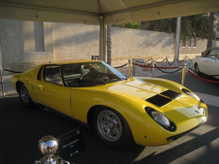 Motor Show anyone? - Page 4 - Middle East - PistonHeads - The image showcases a vintage yellow sports car on display. The car, with its striking yellow body, appears to be a Lamborghini Miura, judging by its distinctive side proportions. The car is parked under a concrete canopy, giving it some protection from the elements. A black sign with "1976 Lamborghini Miura" written in white on it indicates the model and year of this particular vehicle. In the background, additional sports cars and a classic white sedan are visible, suggesting a motoring event or a car museum.