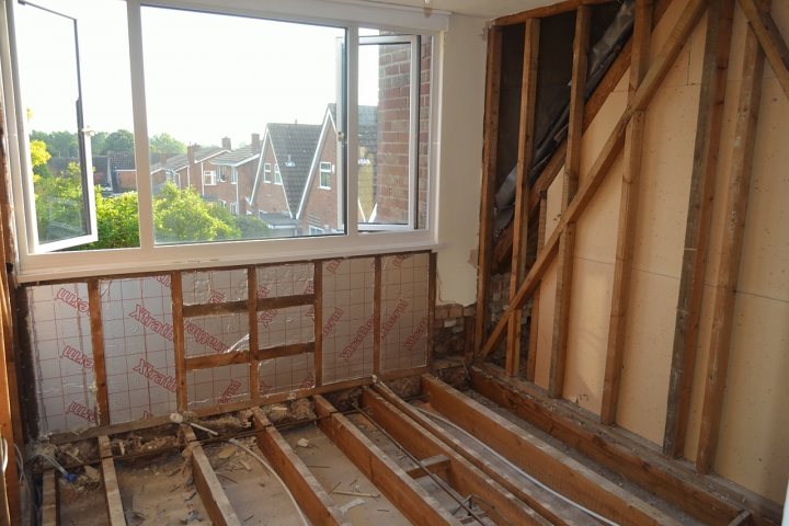 Insulating a dormer house - Page 2 - Homes, Gardens and DIY - PistonHeads - The image depicts an interior view during the construction process. Notably, there is a significant layer of wood which has been removed or is in the process of being removed, exposing the underlying layers. The room is relatively sparse, with just a few pipes visible. The focal point of the image is the large, open window with a view of a brick building outside. The natural light from the window illuminates the wooden floorboards and the beams and studs of the exposed walls. The space appears to be structurally sound but in the midst of a remodeling project.