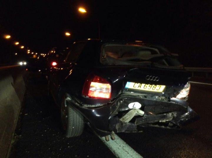 The image depicts a nighttime scene on a busy freeway, illuminated by the headlights of vehicles. The most striking feature of the image is the heavily damaged rear end of a dark-colored sedan. The trunk area is completely destroyed, showcasing the aftermath of a severe collision. Debris and possibly broken glass are scattered around the scene. Additional traffic is passing in both lanes, adding to the sense of the chaos that likely preceded the scene.