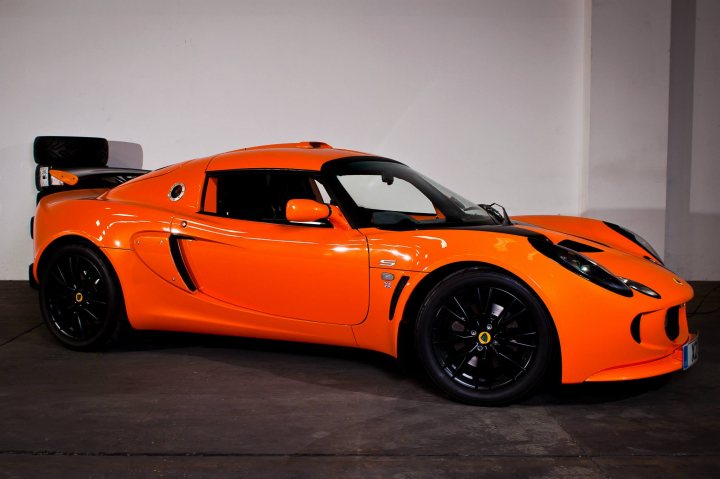 Exige S - Should I? - Page 1 - Elise/Exige/Europa/340R - PistonHeads - In the image, an orange sports car rests in a stark white room. The car's sleek design includes a unibody structure and a cloth roof, giving it a modern and sporty look. The vehicle is positioned at an angle, allowing a clear view of its front and side. Despite being stationary, one could almost imagine the car's potential for speed and agility.