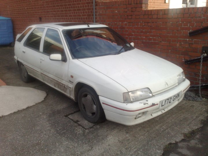 Pistonheads Daily Workhorse Runner - The image shows a white compact car parked on a paved surface alongside a brick wall with what appears to be a black chain or wire attached to a fixture in the wall. The vehicle has a slightly open door on the right and is covered in various spots with dirt or dust, suggesting it is an older model or has been parked for some time. Above the car, a patch of daylight indicates an outdoor setting, likely an urban area. The style of the car suggests it could be from the late 80s to early 90s.