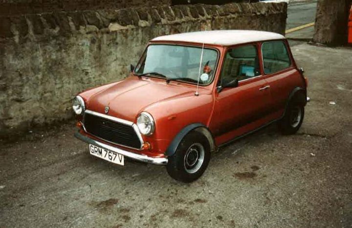 Pictures Of Your Minis! - Page 3 - Readers' Cars - PistonHeads - The image showcases an old-fashioned red and white Mini parked in a paved area. The car is positioned on the driver's side of the road, angled towards the bottom right corner of the photo. It has a single white-walled tire, and unique fender flares cover its tiny black wheels, giving it a distinct retro look. The Mini Cooper badge is prominently visible on the front wheel arch, and the European style license plate is dark-colored, which is typical of European vehicles. The car is parked next to a low stone wall, suggesting a roadside or fence location. The overall scene belongs to a black and white photo, adding a timeless, nostalgic quality to the image.