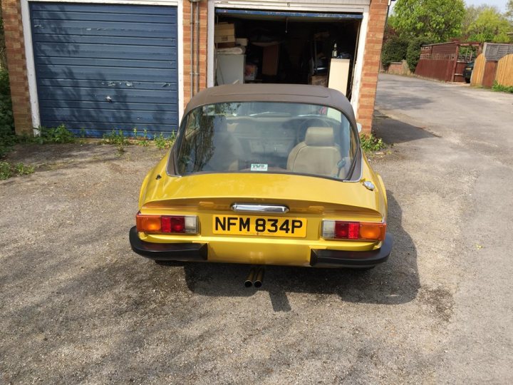 Early TVR Pictures - Page 90 - Classics - PistonHeads - The image features a vintage car, specifically a yellow or gold sports car, with a visible European license plate. It is parked outside an empty garage, which has a closed door and a stepped roof. The ground appears to be a dirt lot, and there are some plants and shrubs in the foreground. The car has its windows down, and it's positioned at a three-quarter angle to the viewer.