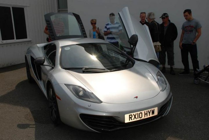 Sunday Supercar Pistonheads - The image showcases a shiny silver sports car parked outdoors. The car, with the hood propped open, is the centerpiece of the scene, drawing attention from several bystanders. These individuals, mostly men, are seen standing or walking around the car, some even carrying handbags. The backdrop to this gathering is a plain white wall, which serves to highlight the car even more. The overall atmosphere suggests an event or display related to the sports car.