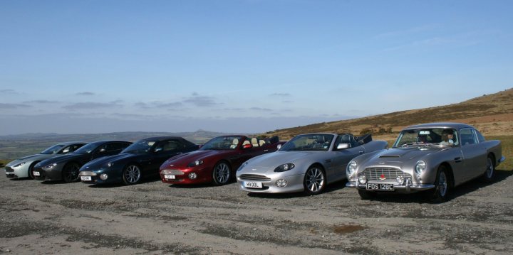 A Georgeous Run Out - Page 1 - Aston Martin - PistonHeads - The image shows a serene scene of a gravel parking lot on a clear day. Five vintage sports cars are parked in a row, each exhibiting a unique color and design that ranges from sleek red to classic silver. The cars are positioned in such a way that their front ends are facing the viewer, revealing the front grilles of their distinct models. In the background, rolling hills rise against a blue sky, adding to the picturesque setting. The license plates on the cars are clearly visible, suggesting that this is a valued collection or event.