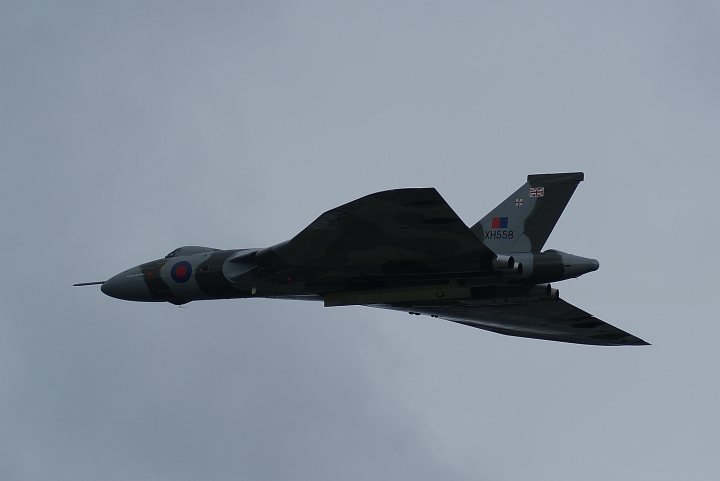 A fighter jet flying through a blue sky - Pistonheads - The image shows a fighter jet soaring high in the sky. It's predominantly clad in a camouflage pattern consisting of hues of gray and green, which blend well with the overcast weather. The jet's tail, visible in the clear blue sky speckled with fluffy white clouds, proudly displays a small British flag, indicating its national affiliation. The jet is captured mid-flight, leaving behind no visible trail from its flight path. The photograph is tinged with darkness, possibly due to overcast weather conditions, and is taken from a perspective that offers a side view of the aircraft.