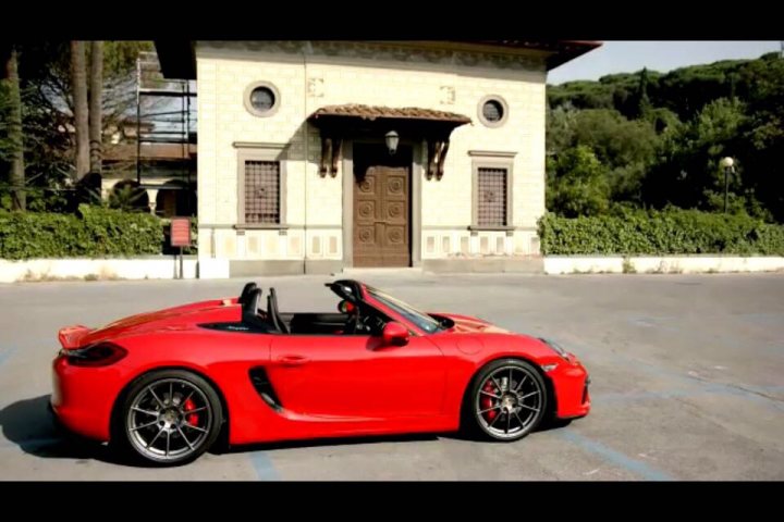 GT4 colours  - Page 52 - Boxster/Cayman - PistonHeads - The image shows a red sports car parked in front of a white building with neoclassical architectural features. The car appears to be a convertible model, possibly a Porsche, given its curved and aerodynamic design. The ground is paved with small stones, and there are green bushes and trees in the background, suggesting a well-maintained environment. The sky is partly cloudy, indicating a lively day outside. The overall setting is picturesque and calm.