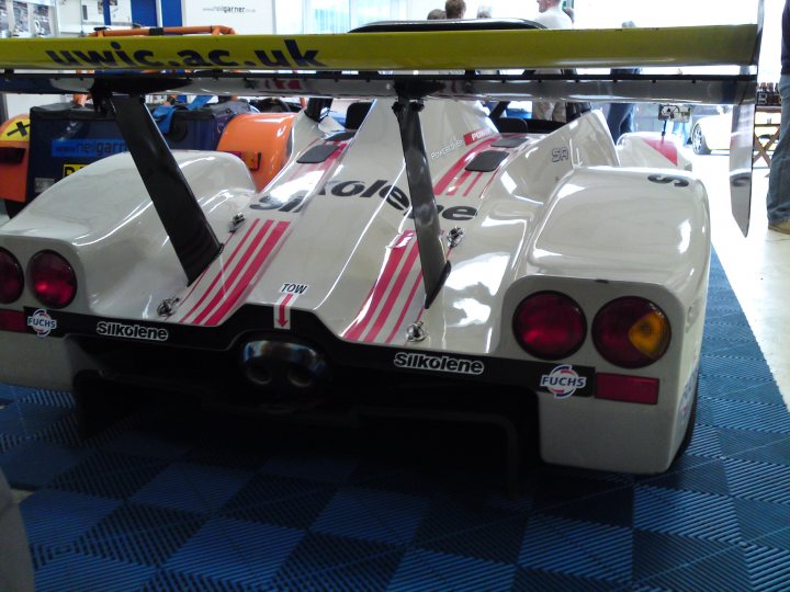 Neil Garner Open Day - Good Friday - Page 1 - South West - PistonHeads - The image captures the rear end of a white and red racing car, parked on a blue and white patterned floor. The car is adorned with several logos, but the text "71 TECH" is particularly prominent on the back wing. The overall color scheme of the car is predominantly white with accents of red stripes. The car's back wing, which is painted black, contrasts with its sleek body. The setting suggests that the car is on display, as there are people in the background, possibly admirers or attendees of an event. The car is positioned in such a way that the license plate is not visible, adding an air of mystery to the scene.