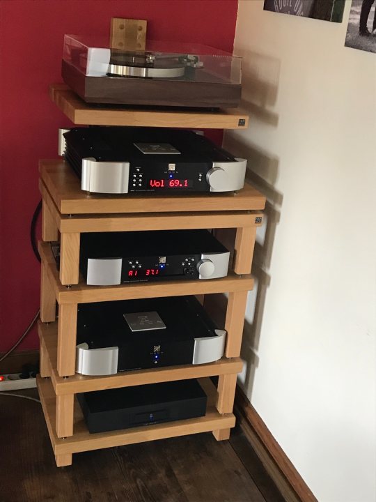 What’s your Hi-Fi set up? spec and pictures please  - Page 20 - Home Cinema & Hi-Fi - PistonHeads - The image depicts a collection of audio equipment arranged on a wooden tiered stand. At the top, there is a record player, while four amplifiers are stacked below it. Each amplifier has various knobs and dials, indicating its complexity and functionality for audio processing. In the background, there's a red wall and part of a person, suggesting that this setup might be in a home or personal space dedicated to music or sound equipment. The presence of the record player at the top indicates an interest in both vinyl records and modern electronic amplification systems.