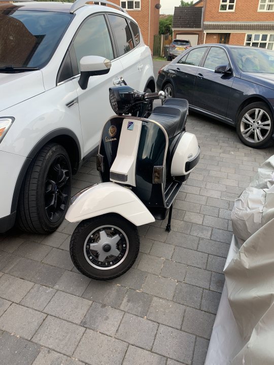 Show us your scooter thread - Page 5 - Biker Banter - PistonHeads UK - The image shows a motor scooter parked in front of a car on a brick pavement. The scooter is black and has a large windshield, a luggage compartment at the rear, and a small sidecar attached. It's parked next to a white SUV with some additional gear secured to the roof. Behind the vehicles is a residential house with white siding. The sky above is clear, suggesting it might be either early morning or late afternoon.