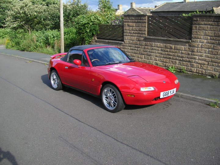 Pistonheads Tallest Driver - The image displays a red sports car parked on a paved street. The car is the primary focus of the image, occupying a significant portion of the scene. It has a shiny exterior, indicative of its likely well-maintained condition. The street on which the car is parked is bordered by a brick wall and plant life, suggesting a suburban or residential area. The sky is overcast, with no visible signs of recent precipitation. The overall ambiance of the image indicates a quiet, possibly early morning or afternoon setting.