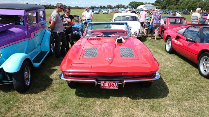 Drive out HalfWay House to North Weald sunday 3rd Aug  - Page 1 - Kent & Essex - PistonHeads - In the heart of a bustling car show, a red classic car, a few others of varying colors and brands, including a blue and pink car and a purple one, dominate the scene. A group of people are gathered around the cars, one of which has a friendly dog on a leash, indicating the jovial atmosphere of the event. Small trees dot the surroundings, and a few tents can be spotted, likely housing vendors or car restorers. The backdrop of the event is filled with more cars, hinting at the wide array of automotive beauty on display.