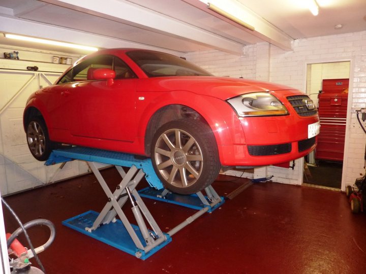 Ramp Buying Pistonheads Garage - The image depicts a vibrant red Audi R8 sitting on a lift in what appears to be a home garage. The lift is a two-post design, which is fitted with a blue pallet. The lift is situated in front of a white garage door and to the side of a window. The surroundings suggest a well-maintained workspace, with tools visible nearby, indicating that this might be a space for regular car maintenance and customization.