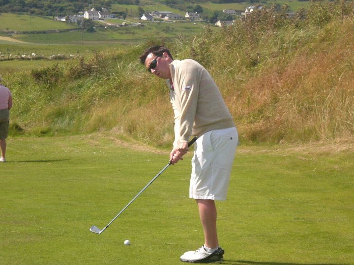 The image depicts a scene from a golf course. At the forefront, a man is preparing to hit a golf ball with his club. He is standing on a lush green lawn, which extends into the distance. The man is wearing a cream-colored sweater and white pants, and he has on sunglasses. In the background, rolling hills and distant buildings can be seen, providing a scenic setting for the man's golfing endeavor.