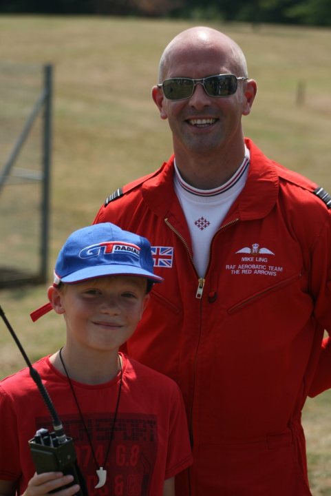 The Red Arrows... - Page 2 - South West - PistonHeads - The image captures a moment of interaction between two individuals. On the left side of the image, a young boy is smiling and holding up a walkie-talkie, perhaps a prop or a souvenir from an event. Oval face, chest, elbows / white smile. On the right, an older man is also smiling and wearing a red flight suit, suggesting a connection to aviation, possibly from a civil or military perspective. The setting seems to be an outdoor area, as indicated by the presence of a metal fence in the background, giving the impression of a sunny day.