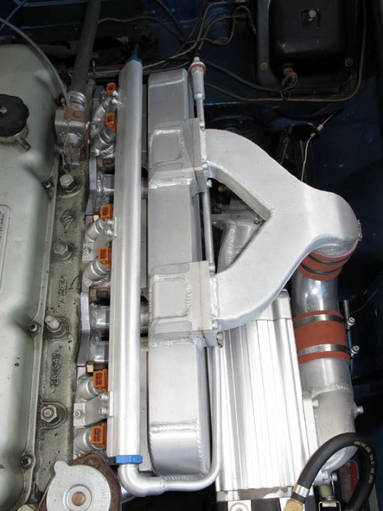 Supercharged Pistonheads - The image shows an engine from under the hood of a convertible vehicle. The primary focus is an aluminum-looking exhaust turbocharger. This turbocharger is accompanied by a cooling system or fan assembly. The engine appears to be worn and dirty, possibly indicating a vehicle with higher mileage. There are several hoses and components visible under the hood, and the overall image provides a view into the complexity and machinery of a car's engine bay.