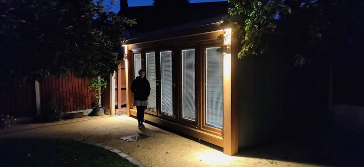 Neighbours built a shed/pub - Page 1 - Homes, Gardens and DIY - PistonHeads UK - The image shows a dark night scene with an outdoor setting. There is a person standing near a wooden structure, possibly a shed or small cabin, which appears to have a slatted roof and is illuminated by a light from within. To the left of the shed, there's a large wooden gate that matches the shed in style and material. The gate has a vertical slat design, allowing visibility through its structure. There is no visible text or branding in the image.