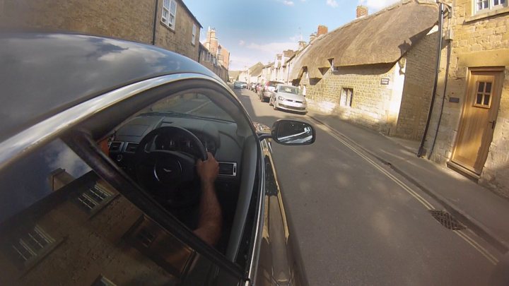 V8 Vantage and GoPro - Page 2 - Aston Martin - PistonHeads - The image shows a view from the driver's seat of a compact car being driven down a narrow street. Time-lapse photography is evident by the line where the buildings appear to be static, indicating movement of the car. The buildings on either side are traditional construction with thatched roofs, suggesting this scene may be in a rural or historical region of Europe. The sky is clear, making for good weather conditions. The car, appearing to be a modern model, has a side rear-view mirror visible in the photo.