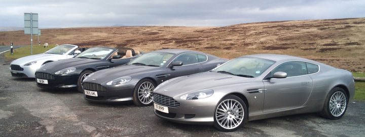 A Georgeous Run Out - Page 1 - Aston Martin - PistonHeads - The image presents a serene scene of four luxury cars neatly lined up in a row on a concrete surface. The cars exhibit a sleek design, with one pair in black and the other in silver. They are parked next to each other, facing the same direction, as if poised for a journey. The backdrop consists of a grassy hill and a fence, adding a rural charm to the setting. The overall arrangement suggests a comparison or showcase of these high-end vehicles.