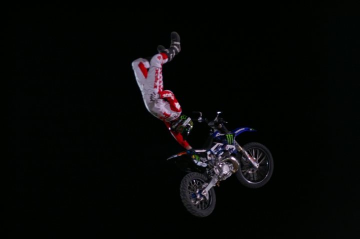 Bull Pistonheads Red Fighters - The image displays a person engaged in motocross, successfully executing an airborne trick on a dirt bike. The rider is clad in a full-bodied, red and white protective suit, which includes a helmet. The motorcycle is airborne, suggesting a jump or obstacle overcame by the rider. The background is a stark black, characteristic of a darkened motocross track at night. The image conveys a sense of skill and daring in the sport of motocross.