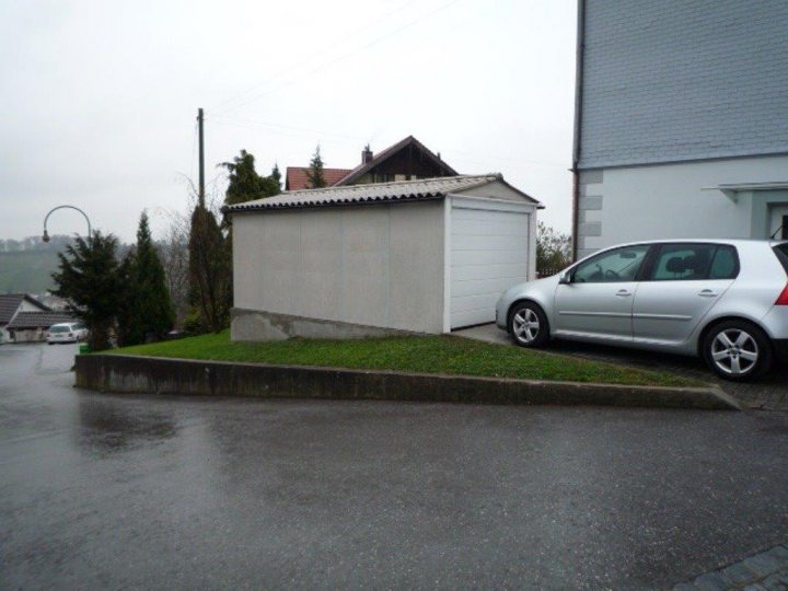 Anyone want a free garage building? - Page 1 - Switzerland - PistonHeads - The image portrays a serene residential scene. Parked on a sloped driveway, a silver car is visible near a white shed or storage unit. The shed, featuring a simple shingled roof and a single door, is set against the backdrop of an elongated grass patch that gently slopes, leading the viewer's gaze towards the car. The setting appears peaceful with a hint of overcast weather, signified by a large gray umbrella situated in the upper left corner of the frame. The absence of people adds a sense of tranquility to the scene.