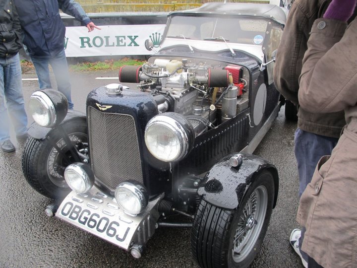 Tax Free Sunday 4th March 2012 - Page 3 - Goodwood Events - PistonHeads - The image showcases a unique, vintage car, possibly a Riley or Bentley, with a distinct sloped front, parked on a wet road. The car has a black and silver color scheme and is likely a convertible due to the absence of a roof. It is part of a track or a similar setting, given the presence of a yellow wall in the background with a Rolex logo and the wet condition of the road. The car is equipped with large headlights and black flags are positioned on the front corners, suggesting it may be on display or participating in a car show.