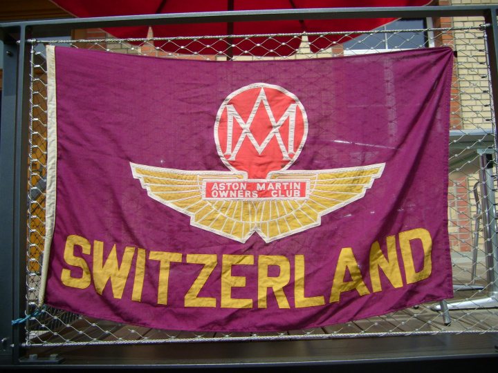 Racer Red Pistonheads - The image showcases a large banner hanging on a metal fence. The banner is predominantly purple with a gold emblem and the word "Switzerland" prominently displayed. There's also a red and yellow logo associated with the country. The text "Aston Martin Owners Club" suggests that this may be a banner from a club or event related to the luxury car brand Aston Martin. The fence on which the banner is mounted is secured by metal bars, providing a pleasing framework for the flag. The backdrop appears to be a building with a red roof, suggesting that the photo was taken outdoors.