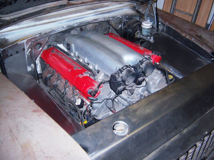 How do you make a 3 door Cosworth go fast????????? - Page 1 - Vipers - PistonHeads - The image shows the engine compartment of a parked vehicle. The engine has multiple components, including a bulky red plastic component positioned towards the center with black plugs on its sides. To the left of this red part, there's a large, metallic engine block with ridged surfaces, and to the right, a smaller black component with numerous openings in a row, which could be a manifold or a similar piece of automotive hardware. The engine bay also features a housing with various tubes leading into the top left corner. The engine compartment appears to be dirty, which might suggest frequent use, oil spills, or work done to the engine, without indicating its overall condition.