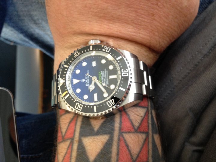 Wrist Check 2015 - Page 1 - Watches - PistonHeads - The image shows a close-up of a person's right arm. On the wrist rests a wristwatch with a blue and black face, displaying the time. The person has a tattoo of black diamond shapes on either side of their wrist. There appears to be a blue laptop display and the lower right corner of a paper or card in the foreground, but the main focus is on the wristwatch and tattoo.