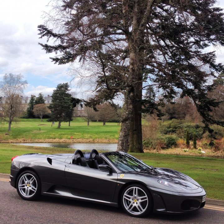 V8V roadster or F430 - tough decision - Page 2 - Aston Martin - PistonHeads - The image shows a sleek, dark-colored sports car parked in front of a grassy area with a body of water. It has a convertible top down, revealing two black seats inside. A person is visible in the background, walking towards the car, adding a dynamic element to the scene. The car is positioned by the side of a parked tree, contributing to a picturesque and serene environment.