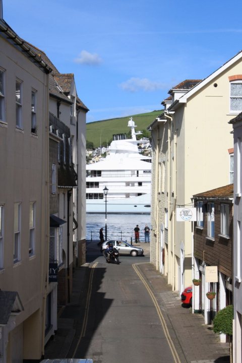 A city street filled with lots of traffic - The image shows a town scene with buildings on either side of a street. In the distance, there is a large yacht close to the shore. A person on a motorcycle is waiting at an intersection, while another individual is walking nearby. The buildings appear to be of European architectural style, and the sky is clear and blue, indicating a sunny day.