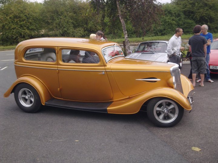 Pistonheads - This image captures a nostalgic street scene, featuring a vibrant gold vintage car that stands out due to its well-maintained condition. The vintage car is parked on the street, with its hood popped open, revealing the engine inside. A group of people are gathered around the car, their attention drawn towards it. Some are clustered closer to the vehicle, while others stand a bit further away. The setting appears to be a typical urban street, with cars parked to the left and trees forming a canopy of green in the background. The image evokes a sense of curiosity and appreciation for classic automotive design.