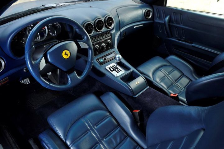 575 miles / colour price sensitive ?  - Page 1 - Ferrari V12 - PistonHeads - This image shows the interior of a vehicle, focusing on the dashboard and seating area. There is a steering wheel prominently in the center foreground, which appears to have a Ferrari emblem. Behind it, the dashboard features a variety of controls and a silver infotainment screen. To the left of the steering wheel, there's an analog clock. The seats are upholstered in a blue material with a diamond pattern, complementing the overall design. The car's interior gives the impression of luxury and high-quality craftsmanship.