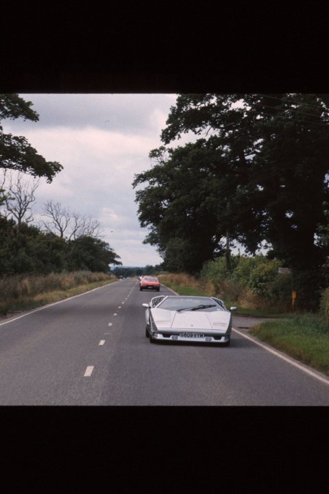 Countach  - Page 14 - Lamborghini Classics - PistonHeads - The image is a still from a color film or video and shows a perspective from the passenger side looking down the road. There are two red cars driving head-on towards the camera, with the first car closer to it. The road appears to be a two-lane highway, and the surrounding environment includes trees and a cloudy sky. The lighting suggests it is daytime and the image likely depicts a landscape scene with vehicles. There are no visible texts or brands in the image.