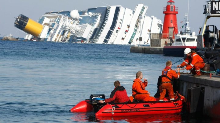 Spot the obvious defect - Page 4 - The Lounge - PistonHeads - The image shows a dramatic scene where a large red lifeboat is being rescued from a capsized ship that's partially submerged in water. The ship, heavily leaning to one side, is a significant part of the image, partially overturned. The lifeboat, filled with people wearing safety gear, is docked alongside the ship's structure. There are at least five individuals visible, some of whom are actively engaged in securing the boat to the dock. The water around the vessels is calm, with no visible signs of currents or strong winds. There is no text present in the image. The image captures the aftermath of an accident, likely involving maritime transportation.