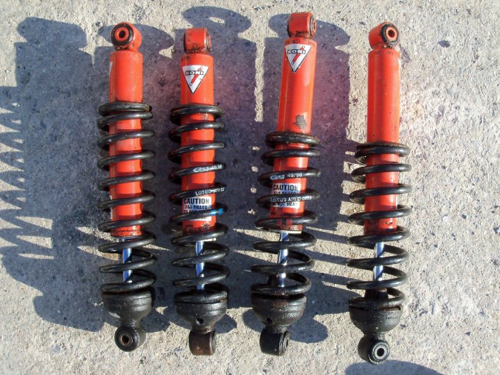 Desperately need to source shock absorbers - Page 1 - Elise/Exige/Europa/340R - PistonHeads - The image shows four red and black shock absorbers laid out on a flat surface, which appears to be a concrete or cement floor. These appear to be used or used-up parts, as indicated by a warning label on each one that states "Vehicle Safety Notice: Do not place this spring inside the vehicle" and includes a hazard symbol. The absorbers have visible springs and connectors, suggesting they were once part of a vehicle, most likely for handling shocks and vibrations to maintain vehicle stability. The warning label seems to be for safety purposes, advising against improper use or disposal.