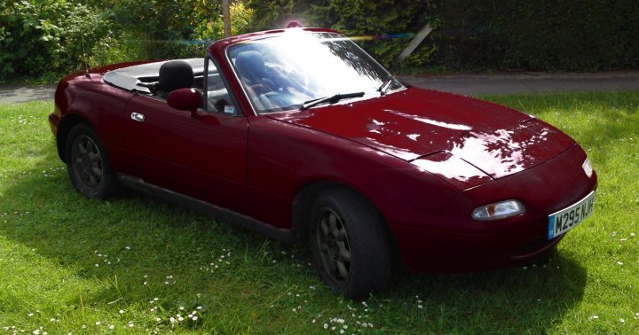 Semisober/insomnia eBay purchase.  - Page 1 - Mazda MX5/Eunos/Miata - PistonHeads - The image shows a small, red roadster under a bright sky, as indicated by the lens flare. The car is parked on grass, slightly angled towards the viewer from the front side. The license plate, although partially visible, contributes to the authentic look and provides context to the location where the car is parked that appears to be in daylight. The setting and the red color of the car suggest that the photo might have been taken during a pleasant countryside drive.