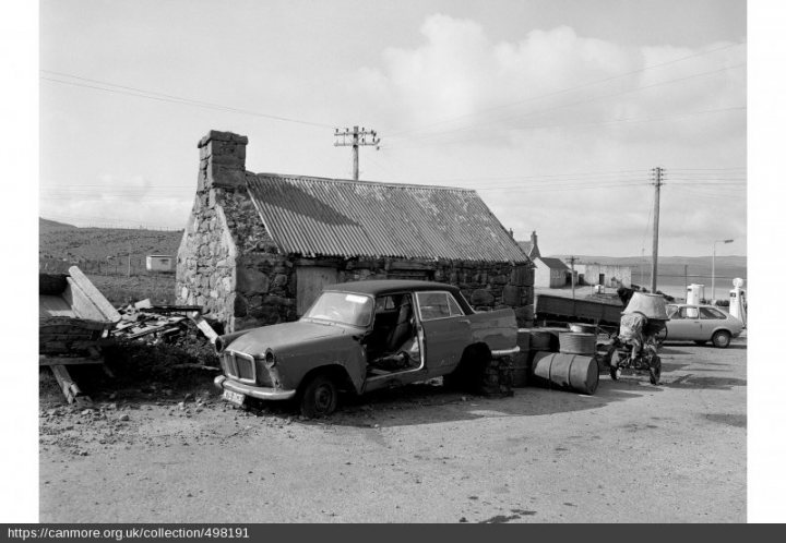 A 'period' classics pictures thread (Mk II) - Page 326 - Classic Cars and Yesterday's Heroes - PistonHeads UK - This is a black and white photograph depicting an old-style building, likely a cottage or shed, with a thatched roof. The structure appears to be in disrepair or abandoned, as indicated by its worn and dilapidated appearance. In the foreground of the image, two vintage cars are parked. These cars have a classic design, reminiscent of early to mid-20th century models. One car is larger and more centrally positioned, while the other is slightly smaller and positioned to the left. The background shows an open landscape with clear skies. The absence of modern vehicles or structures suggests that the location might be rural or in a less urbanized area.