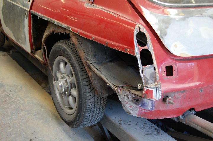 Daylight Pistonheads - The image shows the rear of a vehicle partially taken apart, possibly due to an accident. The back of the vehicle is prominently red and white, displaying visible signs of damage. There is a torn metal panel, possibly from the hood, hanging off the back bumper, along with a dented metal bumper which is still attached. It seems like the vehicle is being prepared for repair or transportation, as it is positioned on a ramp or inclined surface that is often used in repair shops for lifting vehicles. The background is nondescript, suggesting a casual, industrial setting.