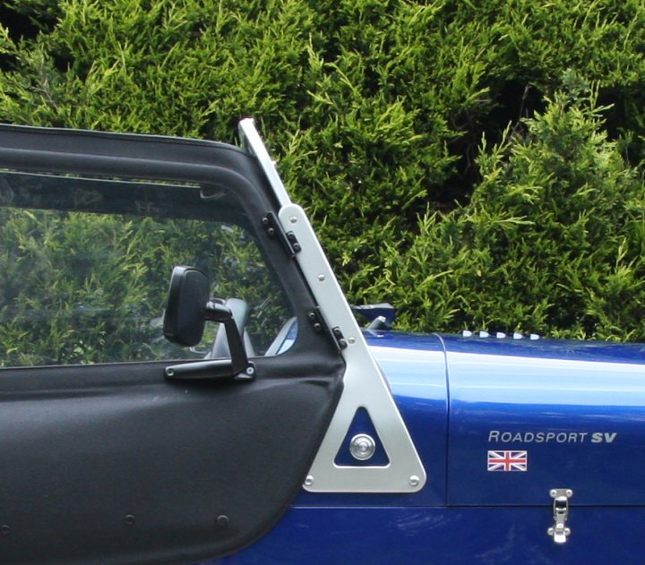 Side Pistonheads Indicator Cycle Repeaters Wings - The image shows a blue vehicle, likely a Sports Utility Vehicle (SUV), set against a background of lush greenery that somewhat obscures the top portion of the vehicle. The vehicle features a side window and passenger handle, which is made of metal and is coated with a black vinyl material for comfort and style. On the side of the vehicle, there's a logo indicating it's a Roadsport SV, a designation that suggests it's a special variant of the Roadsport model. Additionally, there's a small, circular emblem with a British flag, also positioned on the side of the vehicle. The style of the image is a clear, high-resolution photograph that captures the vehicle's details and features.