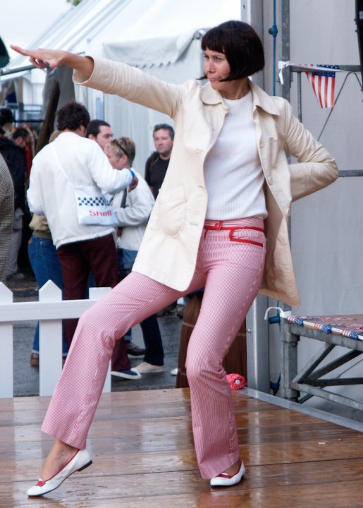 A woman in a white dress holding a tennis racket - Pistonheads - The image captures a lively outdoor scene. At the center of the frame, a woman is energetically performing a dance move on a stage. She's stylishly dressed in a beige trench coat paired with red and white striped trousers, and she's accessorized with white shoes that match her outfit. Her right arm is outstretched, following the graceful lines of her movement.

Surrounding the stage, there are spectators attentively watching the performance. A white picket fence, reminiscent of a tranquil neighborhood, provides a sense of boundary between the performer and the audience.

A blue and white checkered awning adds a touch of whimsy to the scene, suggesting a festive or community event. The awning's checkered pattern contrasts vividly with the surrounding greenery, enhancing the colors in the image.

The woman's dynamic pose combined with the attentive audience creates a sense of pageantry and artistic expression. Overall, this image captures a moment of creative energy in an outdoor setting.