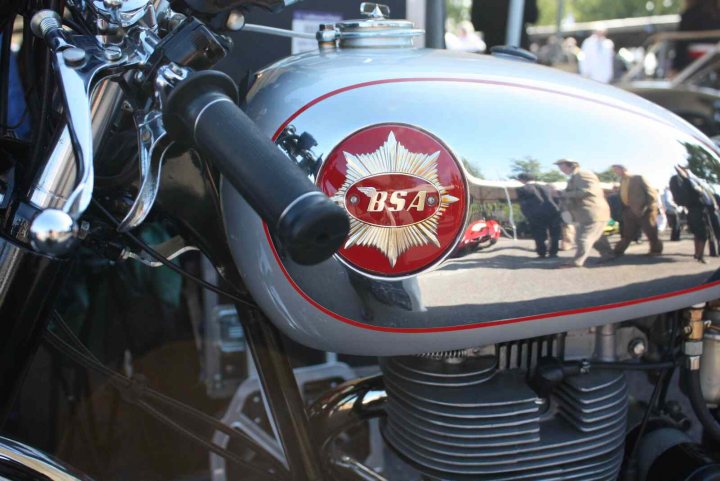 Revival images - Friday  - Page 1 - Goodwood Events - PistonHeads - The image features a section of a motorcycle with a large, well-polished emblem on the gas tank. The emblem includes a shield with a five-pointed star in the center, surrounded by a wreath, and below it, the acronym "BSA1X" is prominently displayed. The motorcycle appears to be in an older condition, with visible scratches and the presence of multiple wires, which may suggest electrical work or a custom build. To the right of the motorcycle, there is a reflection of the sky in the shiny chrome part of the motorcycle, indicating sunlight, and a glimpse of a crowd of people, suggesting that the picture may have been taken at an outdoor event where the motorcycle is showcased.