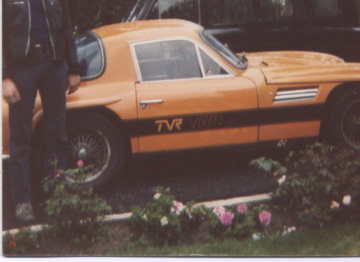 A nice surprise - Page 1 - Classics - PistonHeads - The image is a photograph of a vintage sports car, likely from the 1960s, parked outdoors. The car is predominantly orange with black and silver detailing around its side. There's a hood ornament, but it's not entirely distinguishable. On the side of the car, the badge reads "TVR Vixen," suggesting its make is TVR and its model the Vixen. The setting is a garden, as evidenced by the presence of flowers in the foreground on both the left and right sides. Standing beside the car is a person wearing a dark jacket, partially visible on the left side of the image.